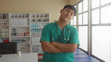 Klinikte kolları çapraz duran, önlük ve steteskop takan genç bir doktor yeterli ışık alan iyi donanımlı bir hastane ortamında kendine güvenini gösteriyor..