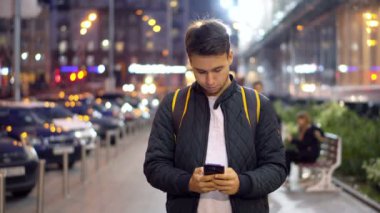 Genç çekici adam yazarak ileti kullanarak Smartphone akşam şehrin bokeh ışık efekti ile sokak.