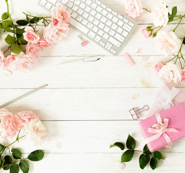 Flat lay women's office desk. Female workspace with keyboard,  flowers pale pink roses,  gifts, accessories on wooden white background. Top view feminine background.Copy space