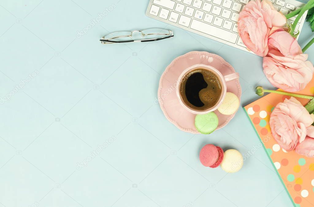 female workspace with keyboard, ranunculus flowers and cup of coffee on blue background 