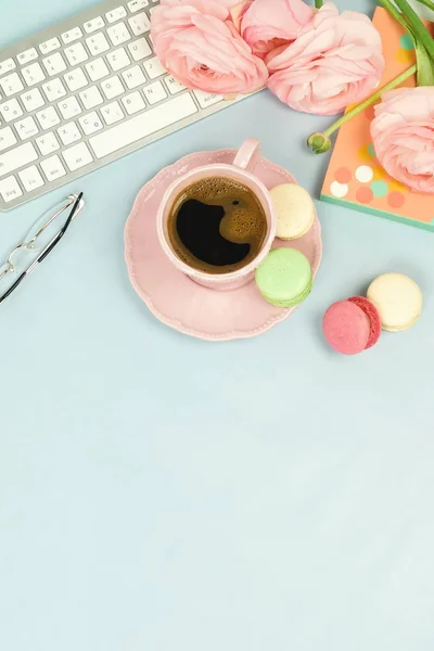 female workspace with keyboard, ranunculus flowers and cup of coffee on blue background