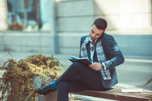 A stylish man is sitting with crossed legs on the bench, talking on his phone and writing in the notebook