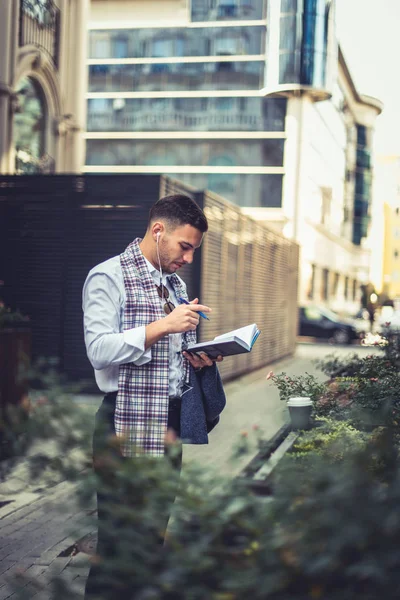 一个自信的商人戴着耳机 拿着一支笔 看着他的笔记本 住在城市的小区里 — 图库照片