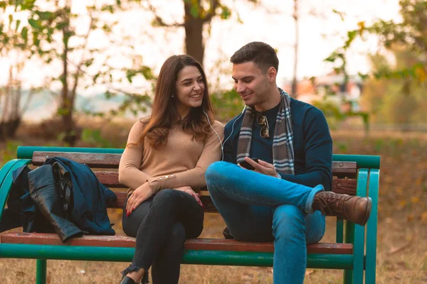 年轻夫妇听着音乐 微笑着 看着电话 坐在公园的长椅上 — 图库照片