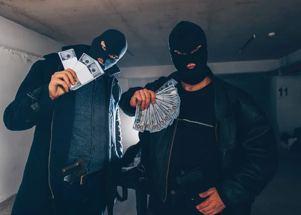 Masked burglars are standing in a dark hallway, holding guns and a bag full of money