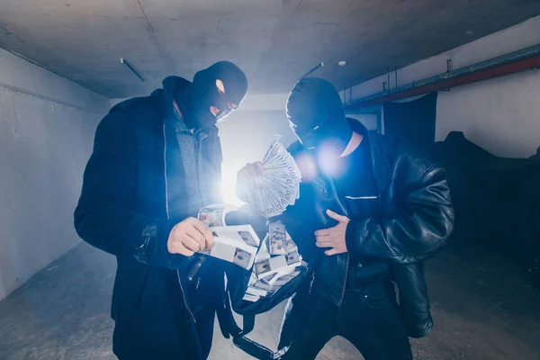 Masked burglars are standing in a dark hallway, holding a bag full of money