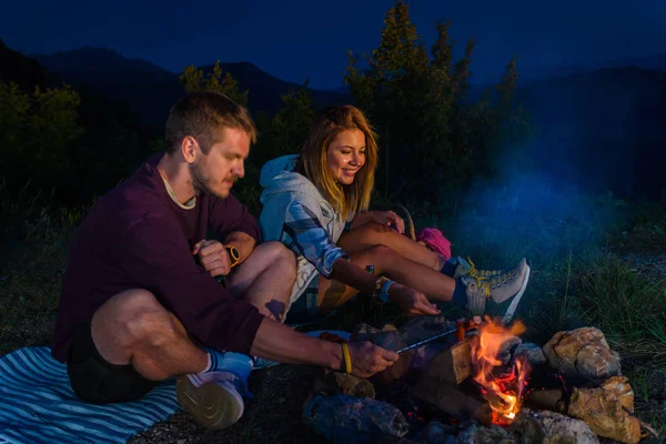 Man and woman baking sausages on the fire, drinking beer and relaxing by the campfire in the mountain top in the night. Romantic campfire concept.