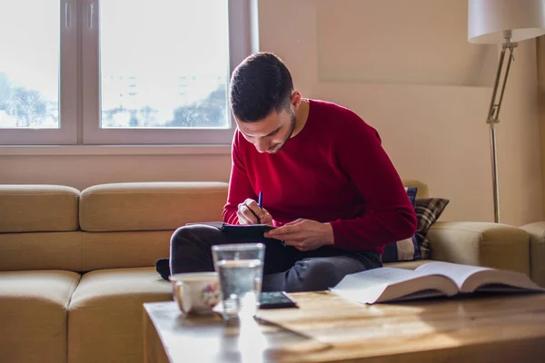 Mann sitzt auf Couch und schreibt zu Hause am Notizbuch — Stockfoto