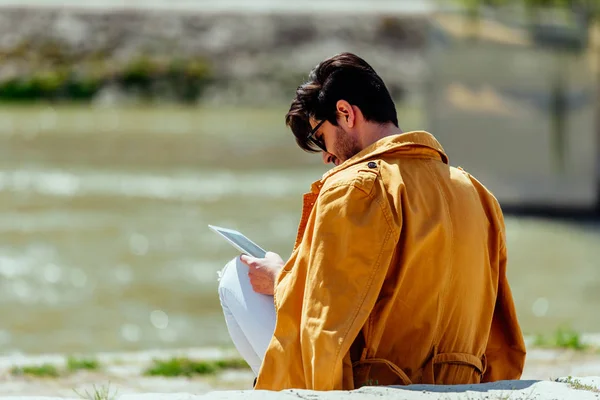 Moderner Typ mit gelbem Mantel arbeitet am Tablet in Flussnähe — Stockfoto