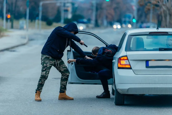 The car thief is pulling the car owner out of his car and trying