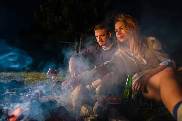 Junges Paar backt Würstchen am Lagerfeuer und trinkt Bier — Stockfoto