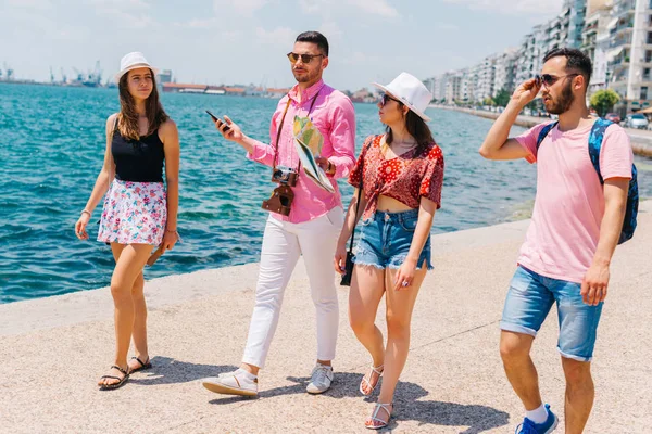 Vier Touristen, die am Meer spazieren, während sie eine Landkarte lesen und — Stockfoto