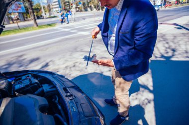 A businessman has parked his car at the side of the boulevard while he checks his car oil, on a sunny busy day. clipart