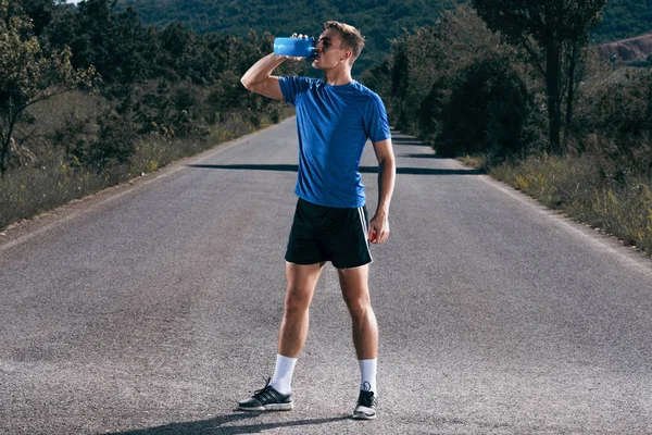 Fit male runner drinking water from a water bottle on an empty r