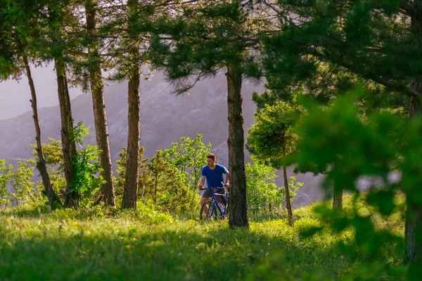 Motociclista de montaña con una camisa azul está empujando su bicicleta a través de —  Fotos de Stock