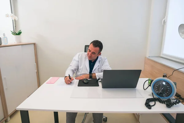 Médico de mediana edad sentado en su despacho y que escribe prescriptio —  Fotos de Stock