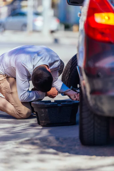 強力なビジネスマンが膝を曲げながら彼の車のフラットタイを変更しようとしています彼のラグレンチで — ストック写真