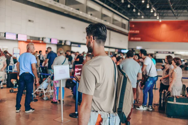 Young man standing with travel suitcase and passport in waiting hall of departure lounge in airport