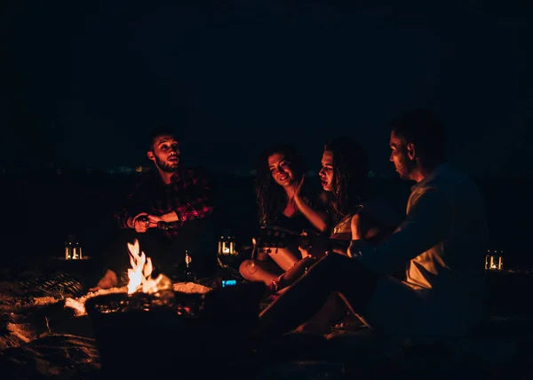 Grupa uśmiechniętych przyjaciół siedzi przy ogniu na plaży — Zdjęcie stockowe