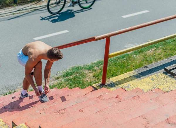 屋外の階段に立って スニーカーの靴ひもを結ぶ運動筋の無気力な男 — ストック写真