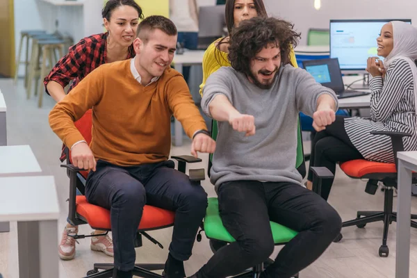 Team building and office fun.Four young cheerful businesspeople in smart casual wear having fun while racing on office chairs and smiling.