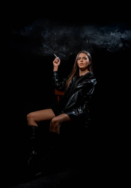迷人的年轻女模特吸烟时穿着时髦的黑色靴子和皮衣 — 图库照片