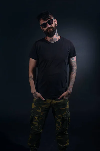 Bearded male model with tattoos and hands in his pocket, dressed in a black t-shirt and sunglasses poses over black background.