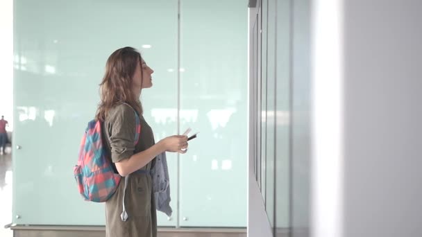 Hermosa joven turista con mochila y llevar en el equipaje en el aeropuerto internacional, cerca del tablero de información de vuelo — Vídeos de Stock