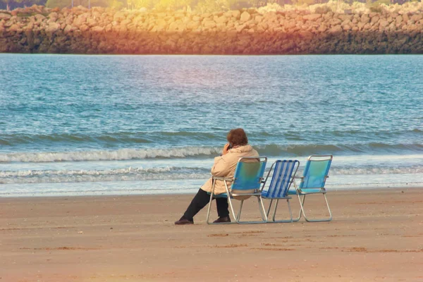 An elderly woman sitting on a chair on the beach and talking on the phone.
