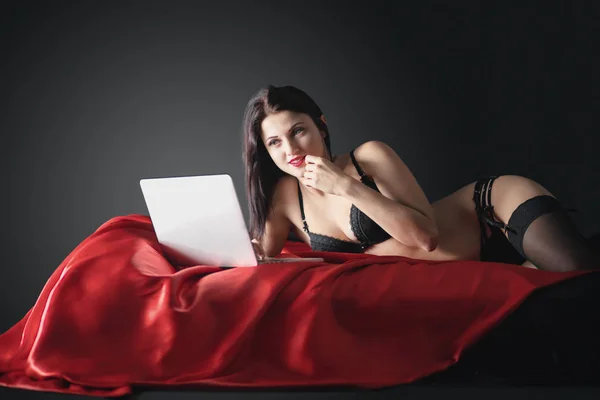 Sexy young brunette woman in black sensual lingerie and in stockings using laptop on red bed in studio.