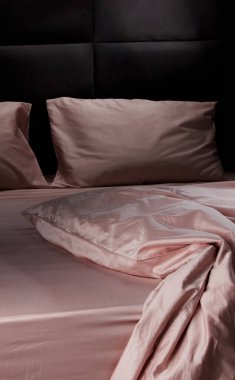 A cozy bed features soft pink sheets and pillows, slightly rumpled, set against a dark wooden headboard. The scene exudes warmth and comfort, ideal for restful sleep.