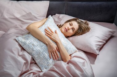 Bir kadın dekoratif bir yastığa sarılıp yatakta uzanırken, yumuşak sabah ışığı yatak odasını doldurur. Düşünceli ve rahat görünüyor, yumuşak renklerde yumuşak çarşaflarla çevrili..