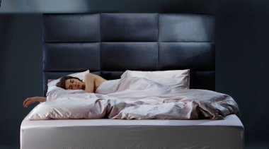 Genç bir yetişkin, yumuşak battaniyelere sarılı olarak huzur içinde yatar. Oda, dinlenmek ve rahatlamak için sakin bir atmosfer yaratan parlak bir yatak başlığıyla minimalist bir tasarıma sahip..