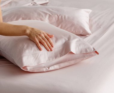 Bir el yumuşak pembe bir yatak düzeninde yumuşak bir yastık kılıfını nazikçe geri çeker. Gündüz vakti modern bir yatak odası ortamında davetkar bir atmosfer yaratır..