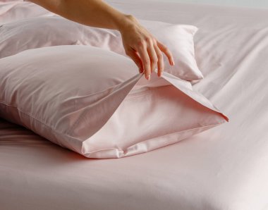 Bir el yumuşak pembe bir yatak düzeninde yumuşak bir yastık kılıfını nazikçe geri çeker. Gündüz vakti modern bir yatak odası ortamında davetkar bir atmosfer yaratır..