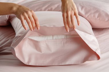 Bir el yumuşak pembe bir yatak düzeninde yumuşak bir yastık kılıfını nazikçe geri çeker. Gündüz vakti modern bir yatak odası ortamında davetkar bir atmosfer yaratır..