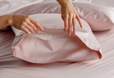 Bir el yumuşak pembe bir yatak düzeninde yumuşak bir yastık kılıfını nazikçe geri çeker. Gündüz vakti modern bir yatak odası ortamında davetkar bir atmosfer yaratır..