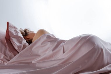 Bir kadın, ipeksi pembe çarşafların altına kıvrılmış, rahat bir yatakta dinleniyor. Güneş ışığı pencereden akarak odada sakin ve sakin bir atmosfer yaratır..
