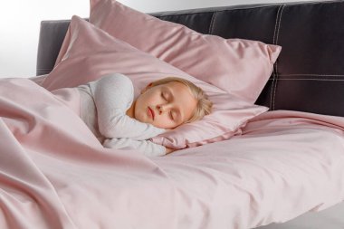 Küçük bir çocuk, rahat bir yatakta yumuşak pembe bir yatağa sarılı olarak huzur içinde uyuyor. Sabah ışığı odayı hafifçe aydınlatıyor, sakin bir atmosfer yaratıyor..