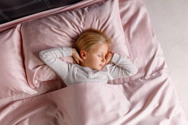 Küçük bir çocuk, rahat bir yatakta yumuşak pembe bir yatağa sarılı olarak huzur içinde uyuyor. Sabah ışığı odayı hafifçe aydınlatıyor, sakin bir atmosfer yaratıyor..
