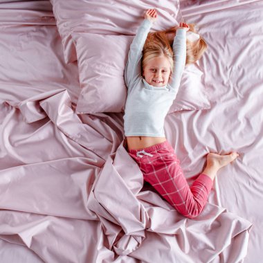 Genç bir kız soluk pembe bir yatakta sessizce yatıyor, saçları yastıklara yayılmış. Düşünceli görünüyor. Yumuşak öğleden sonra ışığı odayı aydınlatıyor. Huzurlu bir atmosfer yaratıyor..