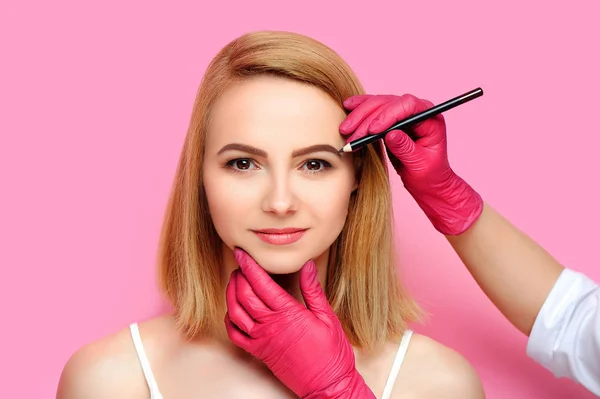 Beauty specialist correcting eyebrows shape with a brown cosmetic pencil against pink background