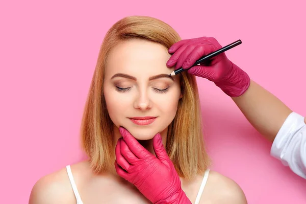 Eyebrows correcting procedure with a cosmetic pencil against pink background