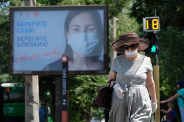 Almaty / Kazakistan - 07.13.2020: İnsanlar trafik ışığı ve maske takmak ve mesafeyi korumakla ilgili uyarı içeren bir Billboard ile kesişme noktasından geçiyor.