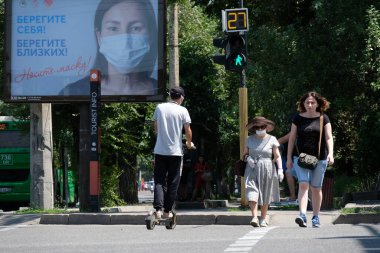Almaty / Kazakistan - 07.13.2020: İnsanlar trafik ışığı ve maske takmak ve mesafeyi korumakla ilgili uyarı içeren bir Billboard ile kesişme noktasından geçiyor.