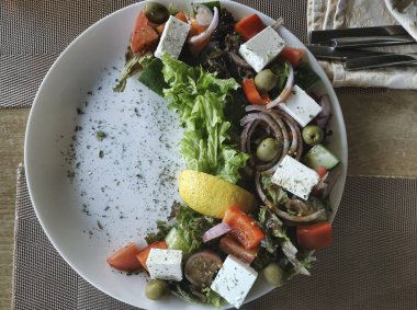 Yunan salatası beyaz bir tabakta hilal şeklinde sunuluyor. İçindekiler: domates, salatalık, dolma biber, kırmızı soğan, çiğ feta, limon suyu, çekirdeksiz zeytinyağı, zeytinyağı..