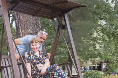 Yetişkin bir kadın bir bankta oturuyor ve arkasında bir yaz akşamı parkında bir adam.