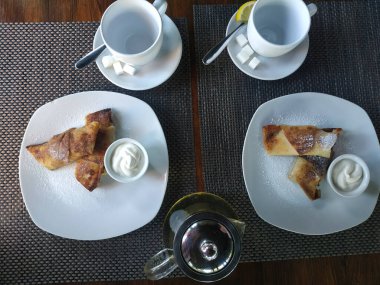 Krepler süzme peynirli ve ekşi kremalı. Masada iki porsiyon, çay için iki boş bardak ve bir çaydanlık çay.