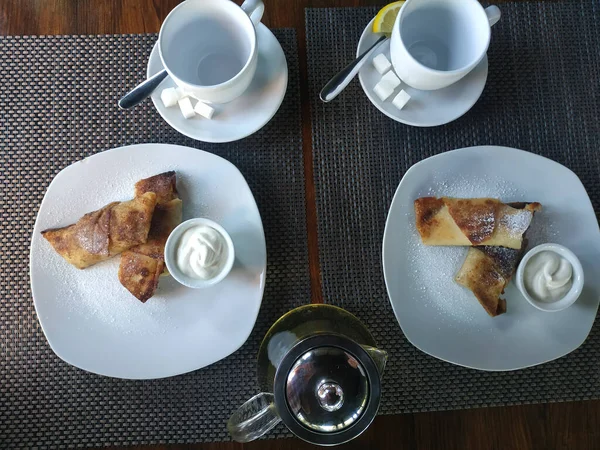 Krepler süzme peynirli ve ekşi kremalı. Masada iki porsiyon, çay için iki boş bardak ve bir çaydanlık çay.