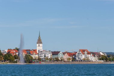FRIEDRICHSHAVEN, GERMANY - 6 Eylül 2025: Güneşli bir yaz gününde açık mavi bir gökyüzünün altında Friedrichshafen, Almanya 'da bir göl havuzu ve bir kilise kulesinin manzarası